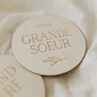 Plaque ronde en bois clair gravée « Future Grande Soeur » avec motif feuille sur tissu beige