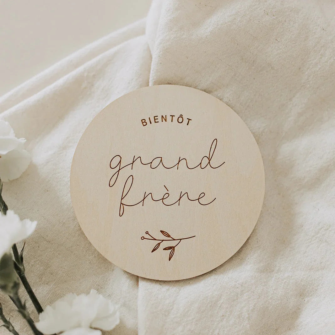 Disque en bois rond gravé « bientôt grand frère » sur tissu beige avec fleurs blanches