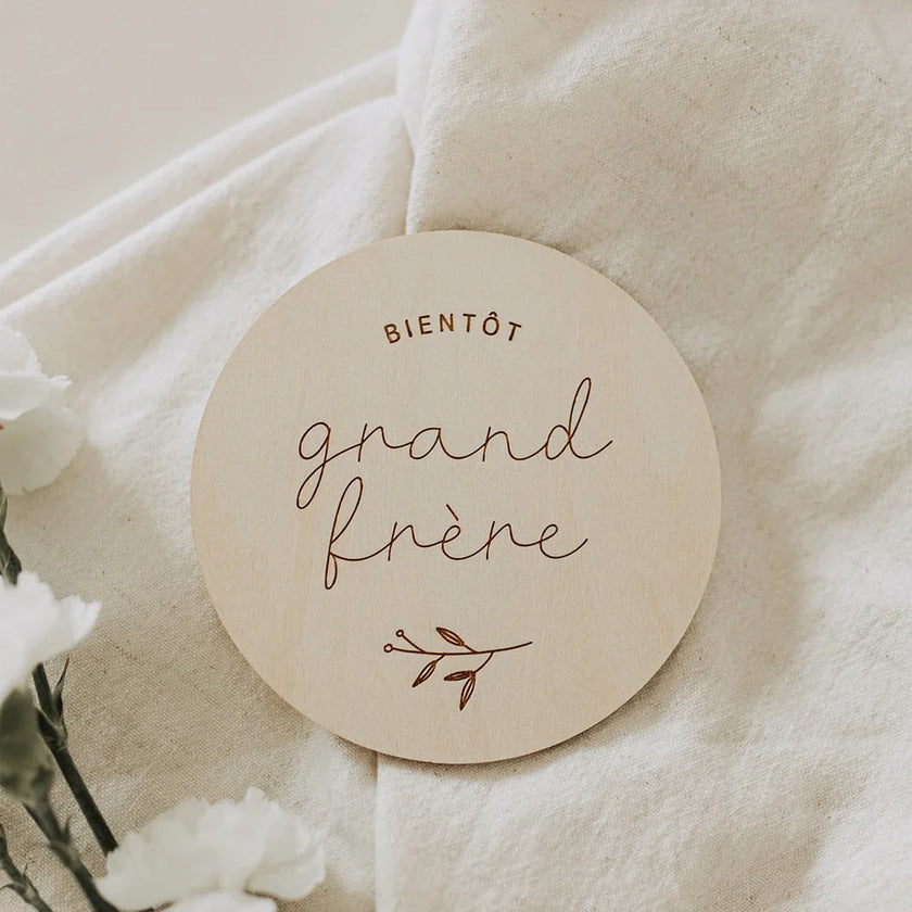Disque en bois rond gravé « bientôt grand frère » sur tissu beige avec fleurs blanches