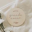 Disque en bois gravé « Bientôt grande sœur » sur tissu beige avec fleurs blanches