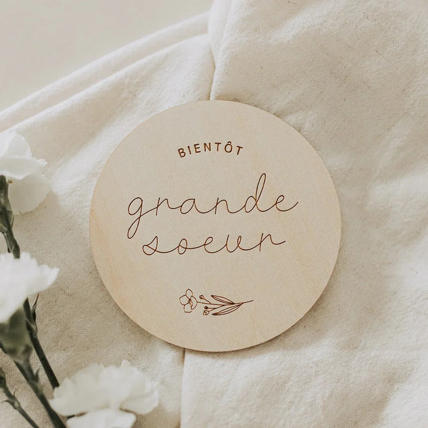 Disque en bois gravé « Bientôt grande sœur » sur tissu beige avec fleurs blanches
