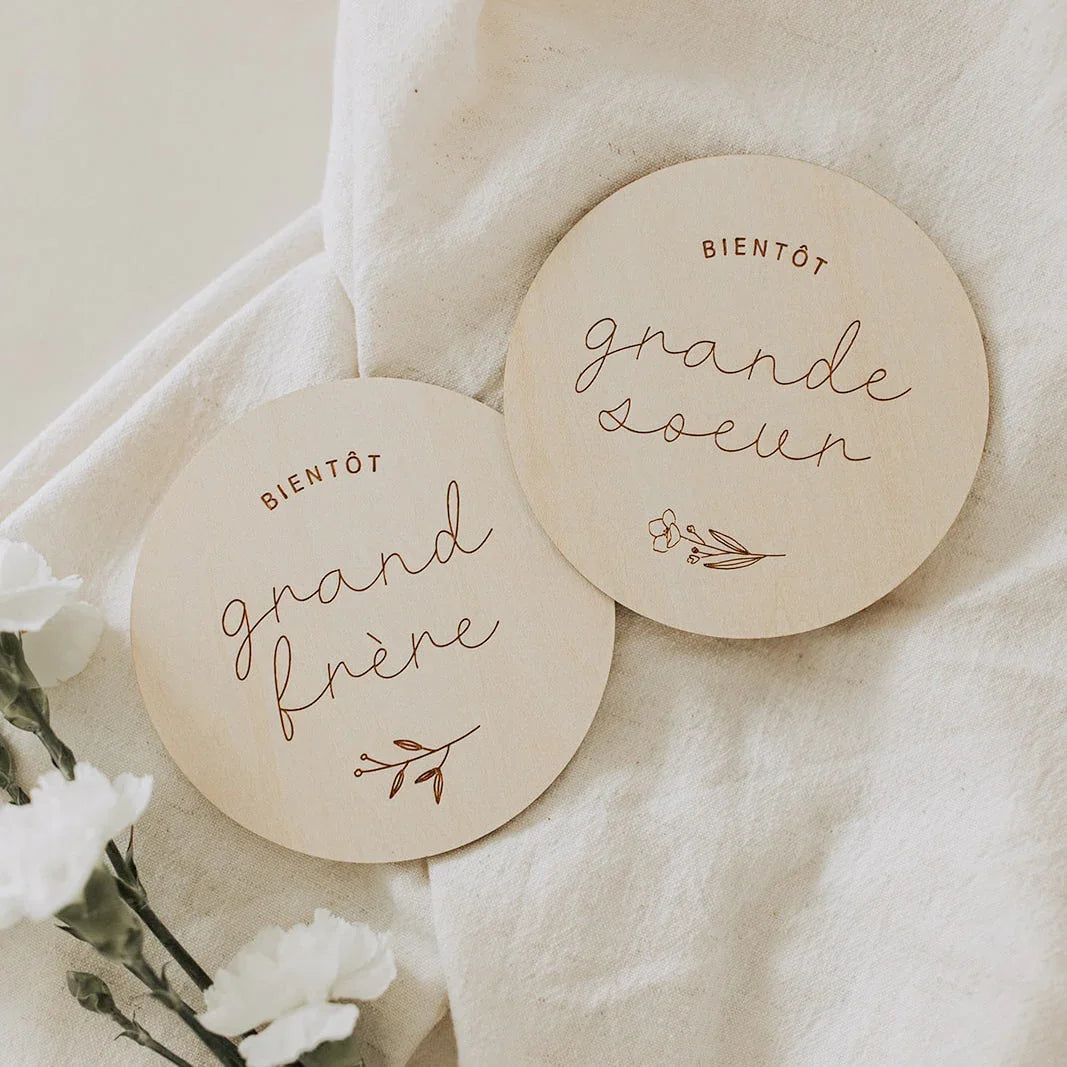 Deux plaques rondes en bois gravées avec inscriptions « Bientôt grand frère » et « Bientôt grande sœur » sur tissu beige