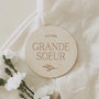 Badge rond en bois gravé "Future Grande Soeur" avec branche décorative sur tissu blanc et fleurs blanches