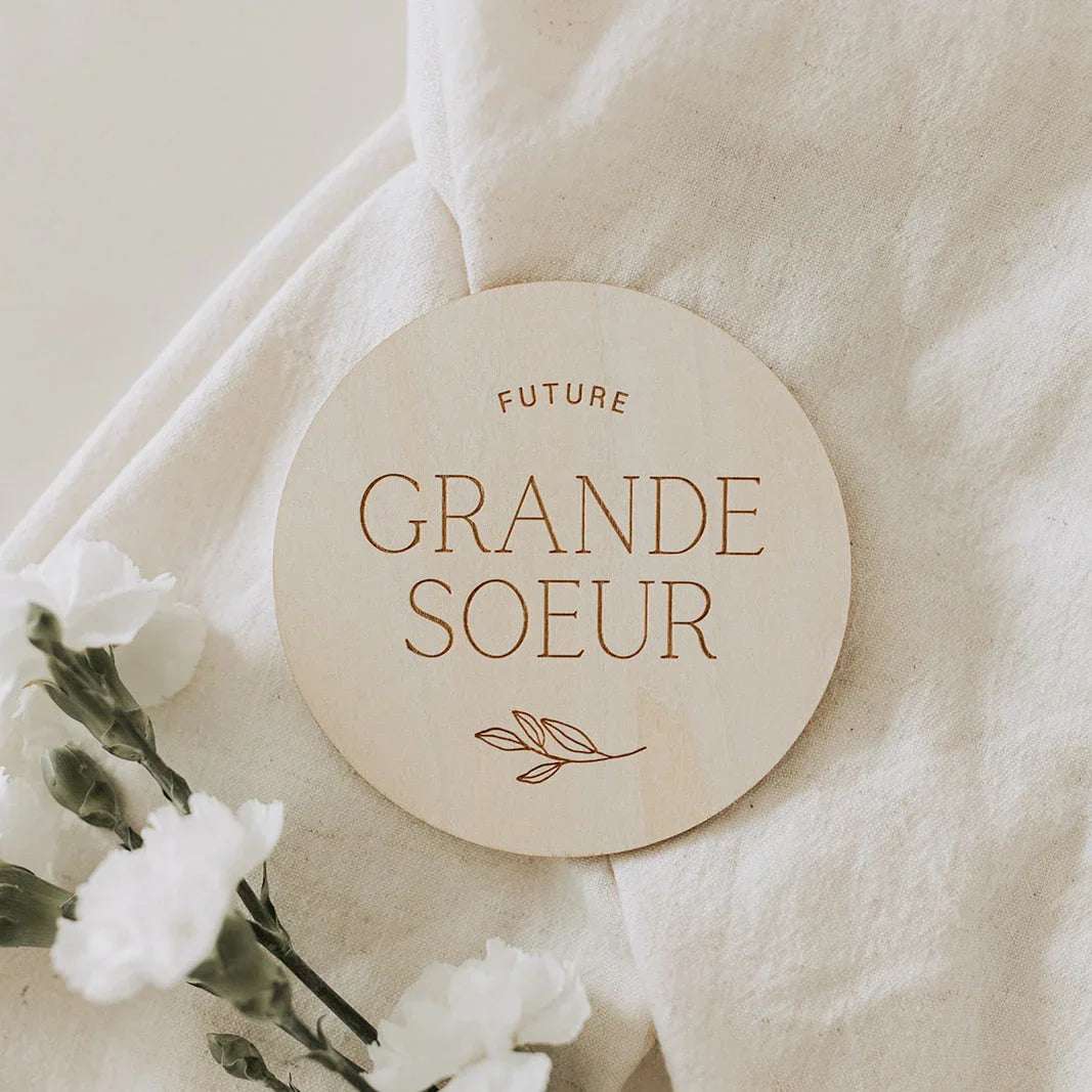 Badge rond en bois gravé "Future Grande Soeur" avec branche décorative sur tissu blanc et fleurs blanches