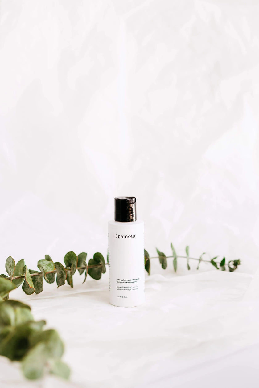 Flacon blanc de liniment oléo-calcaire Énamour avec bouchon noir sur fond blanc et branche de feuilles vertes