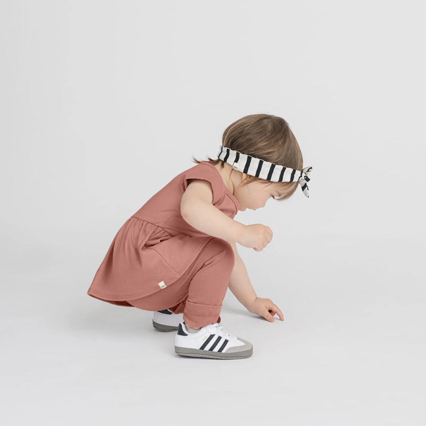 Bébé accroupi portant une robe-pantalon argile, bandeau rayé noir et blanc, chaussures blanches Adidas