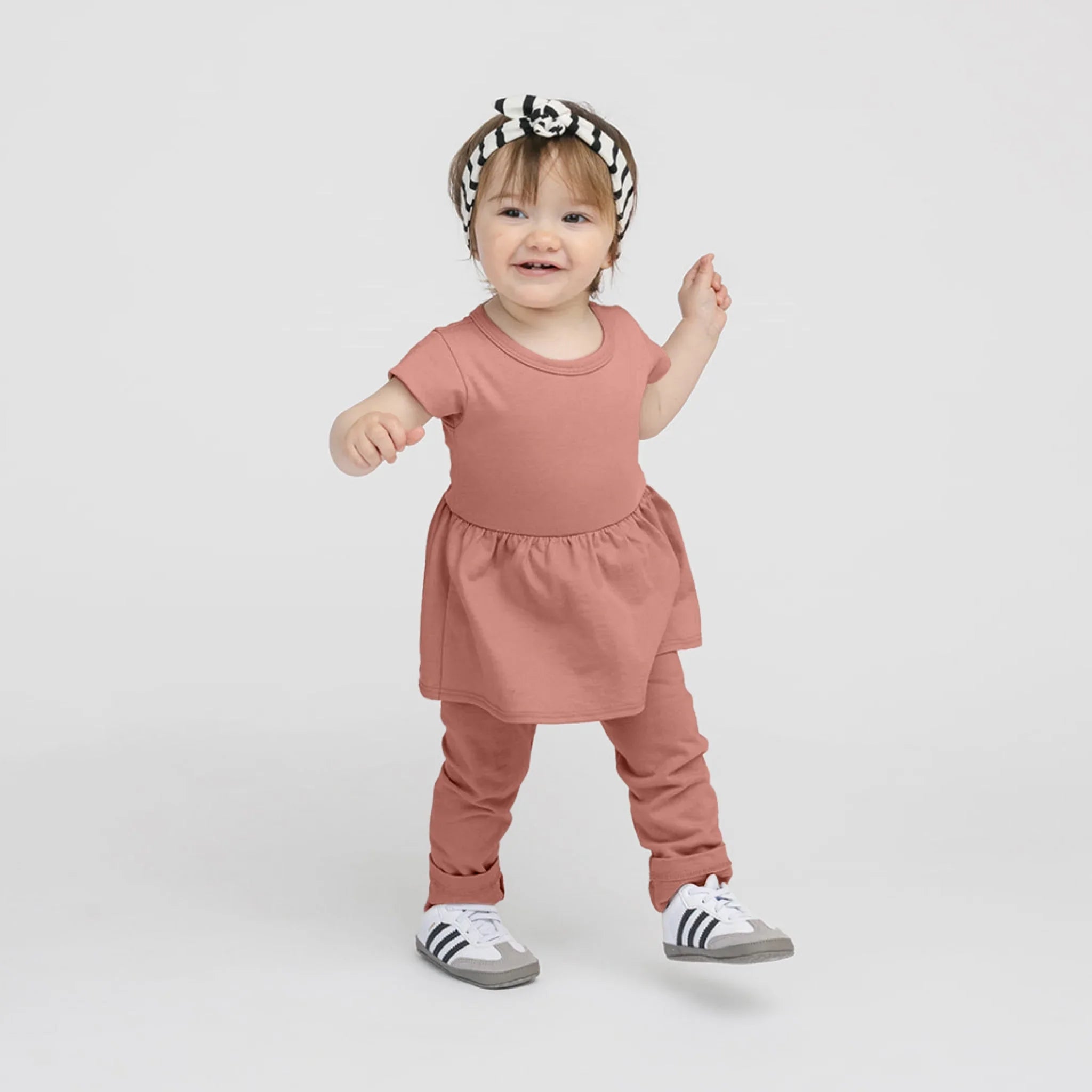 Bébé fille souriante en robe-pantalon argile avec bandeau rayé noir et blanc et baskets