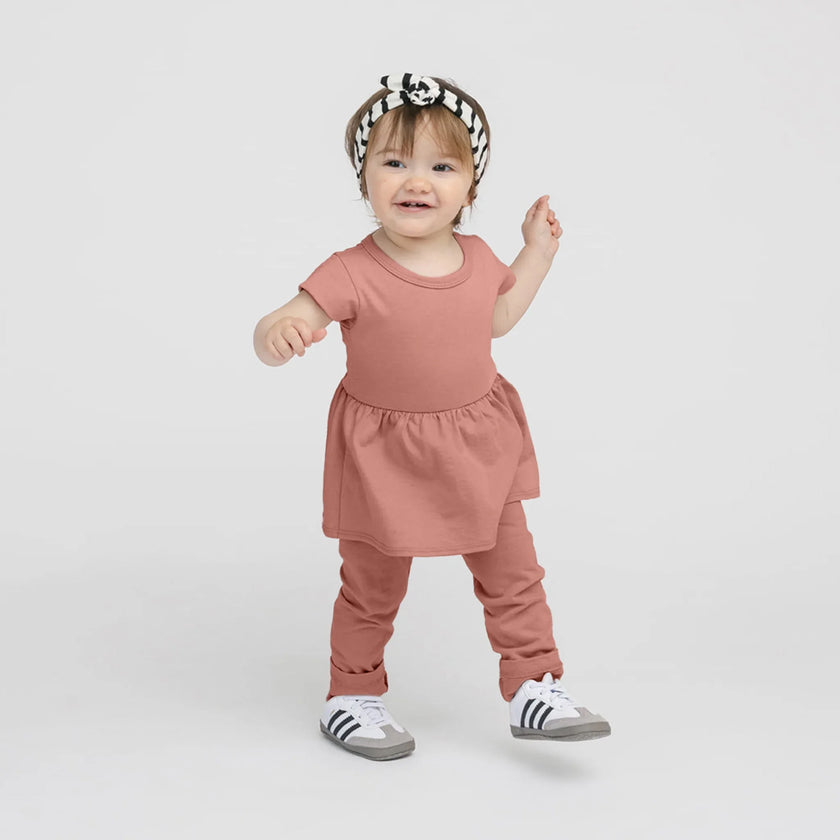 Bébé fille souriante en robe-pantalon argile avec bandeau rayé noir et blanc et baskets