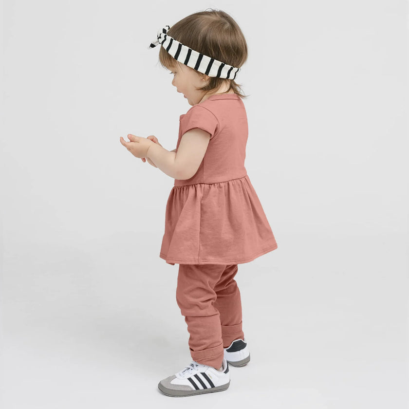 Bébé fille de profil portant un ensemble saumon avec robe et leggings, bandeau rayé noir et blanc, chaussures blanches
