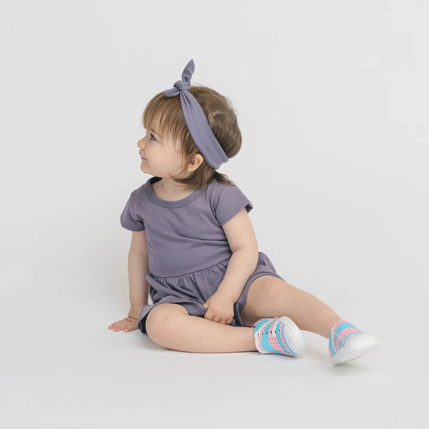 Bébé assise en robe vintage prune et bandeau assorti, chaussures colorées bleu et rose